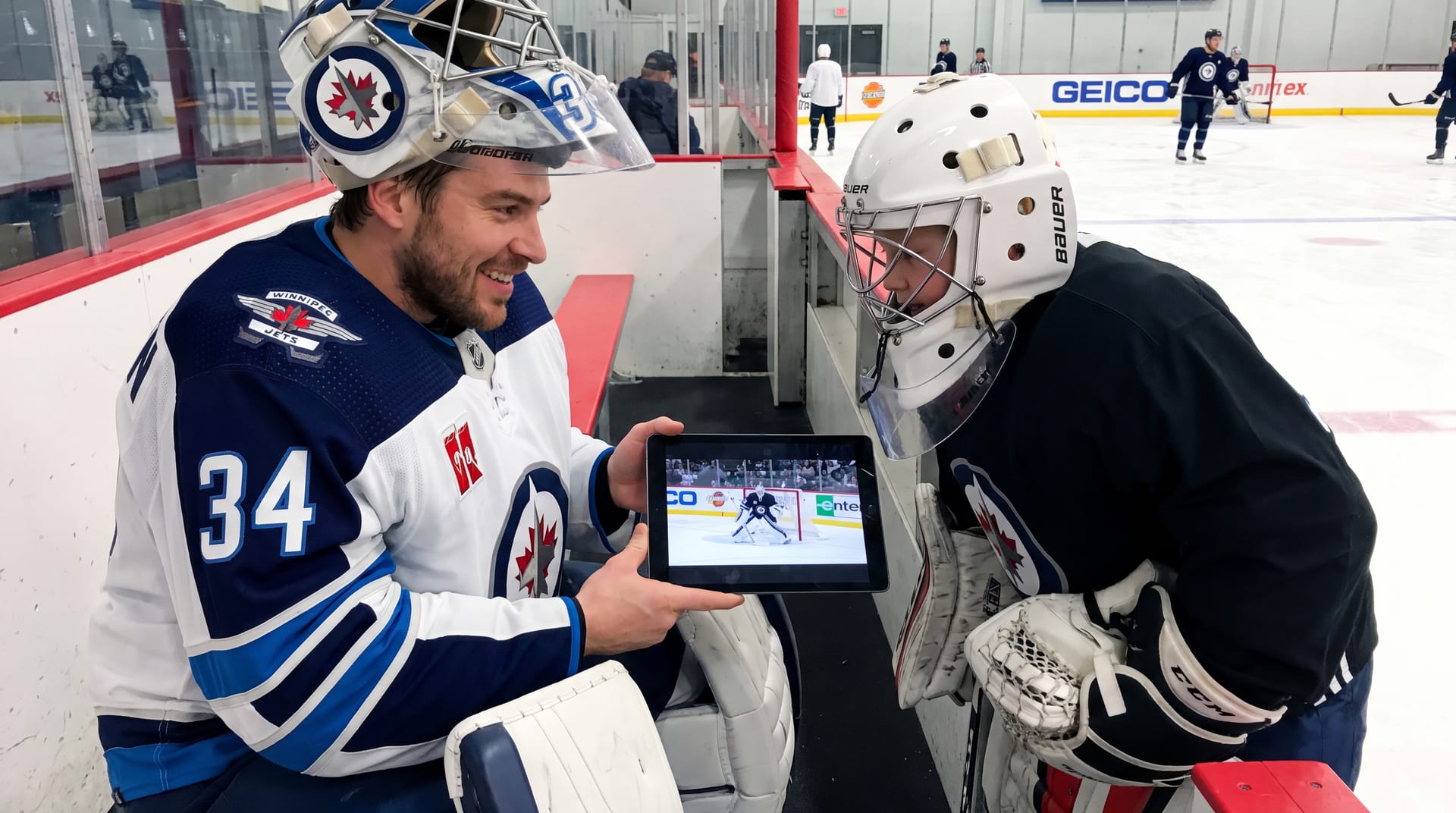 Michael Hutchinson in Winnipeg Jets gear at the bench, showing a young goalie game footage on a tablet